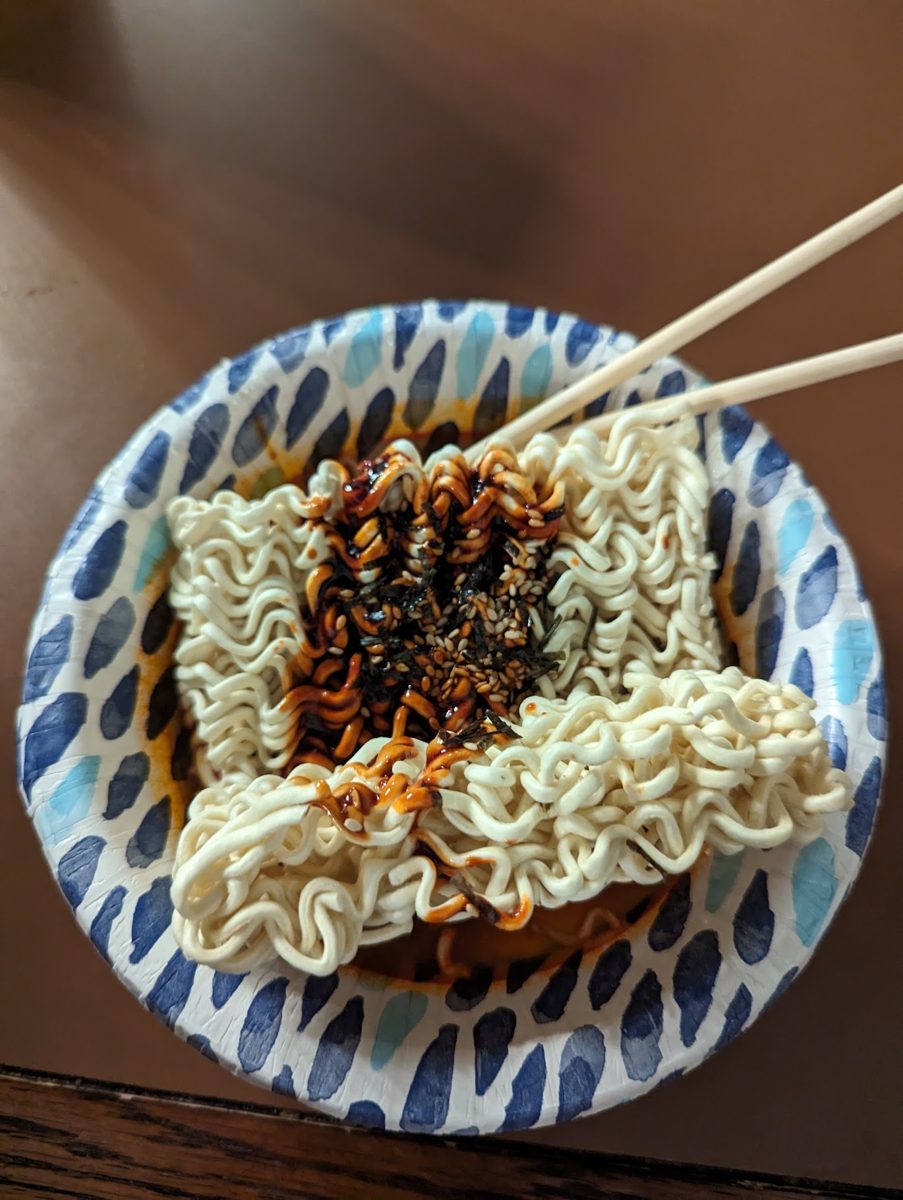 A paper bowl of the spiciest buldak noodles, covered in dark chili sauce, with chopsticks